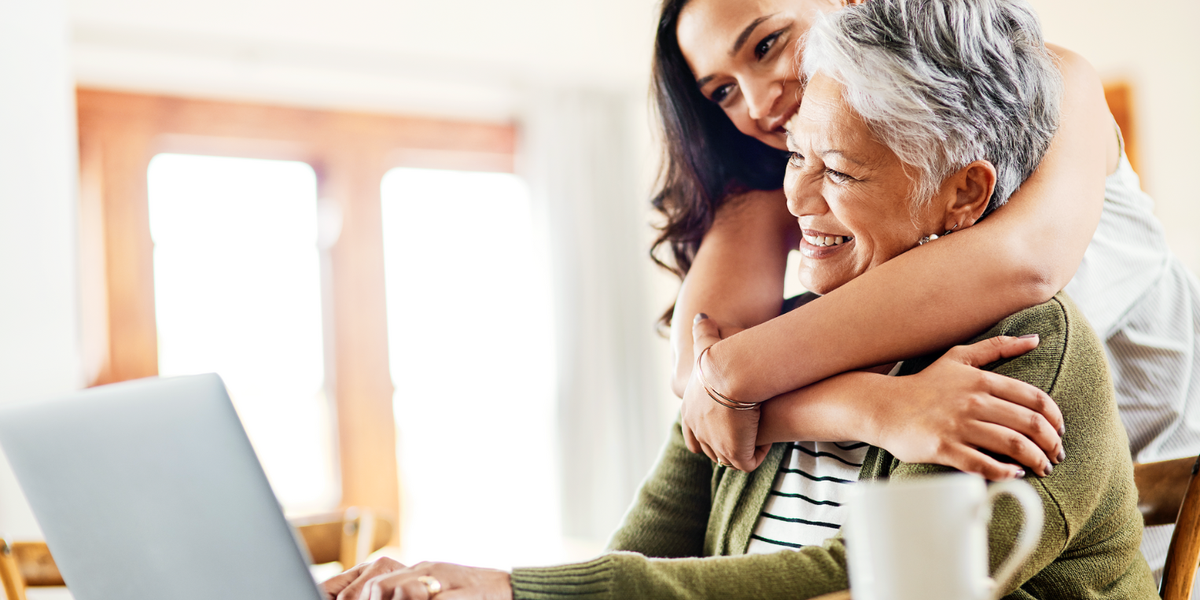 Older woman smiling at a table on her laptop with a younger woman hugging her.
