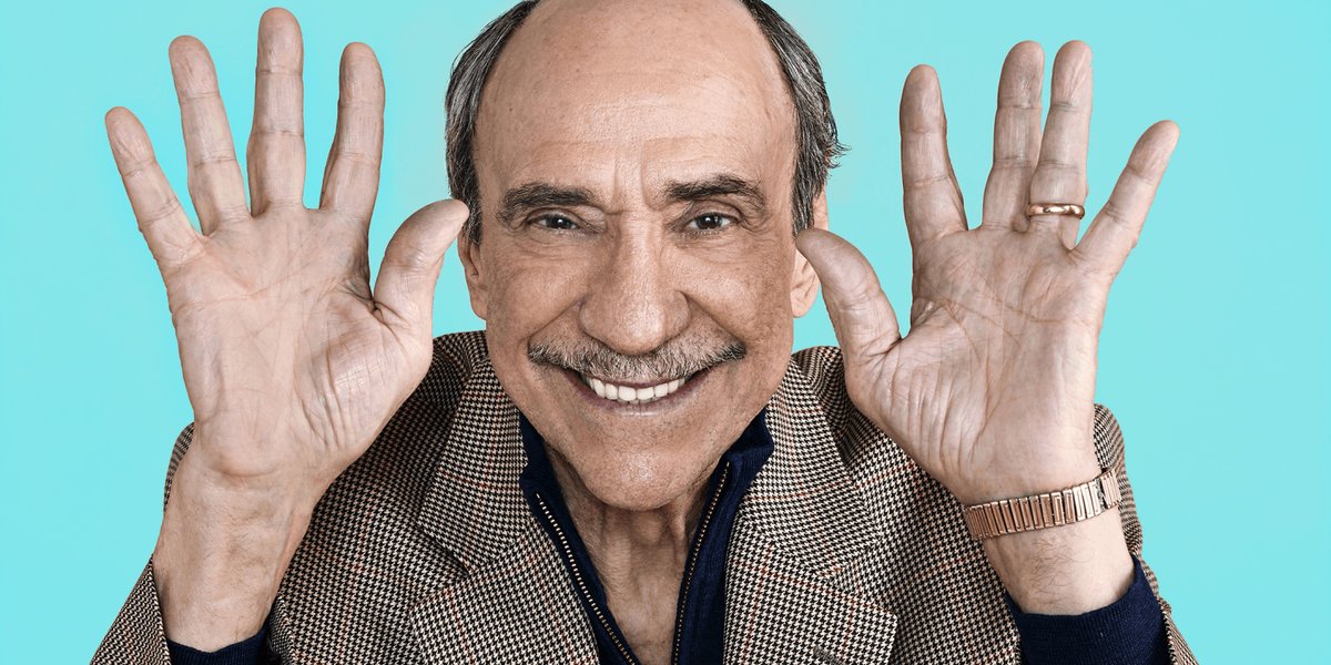 F. Murray Abraham standing against a blue background holding his hands up near his face