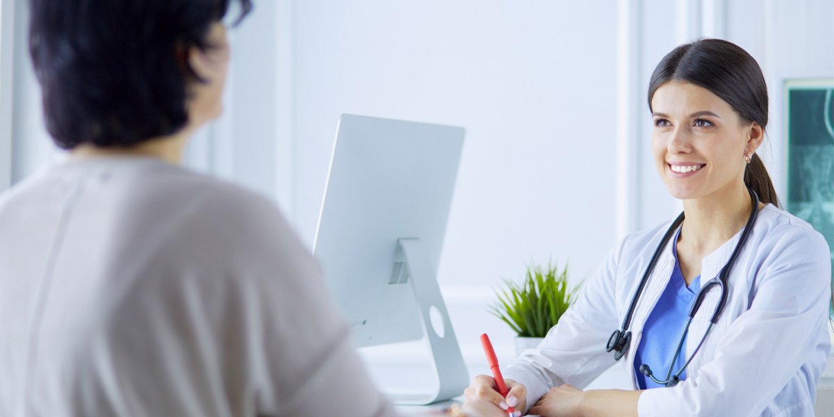Doctor in a white lab coat and blue scrubs speaking to a patient in a grey sweater at a doctor's office while writing down notes