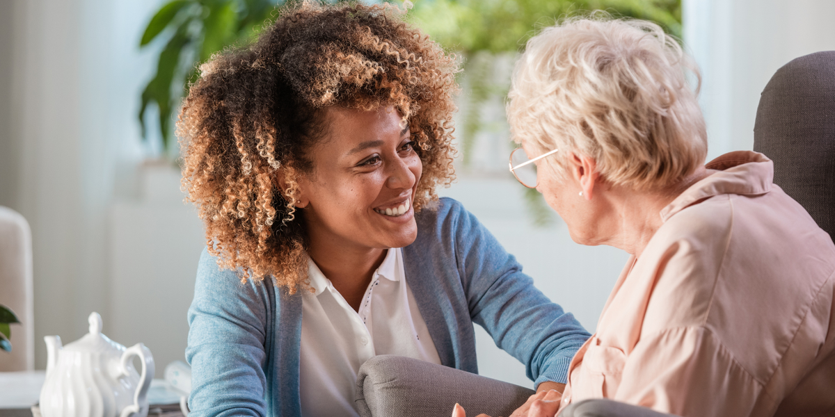 Smiling female caregiver in a blue cardigan holding hands and talking with senior woman in living room