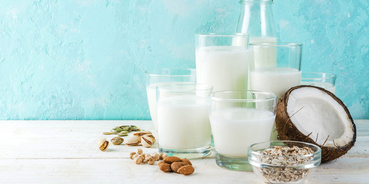 Variety of alternative milks in glasses on a white table in front of a blue background with small bowls of soy, almond, flax, oats, and half of a coconut.