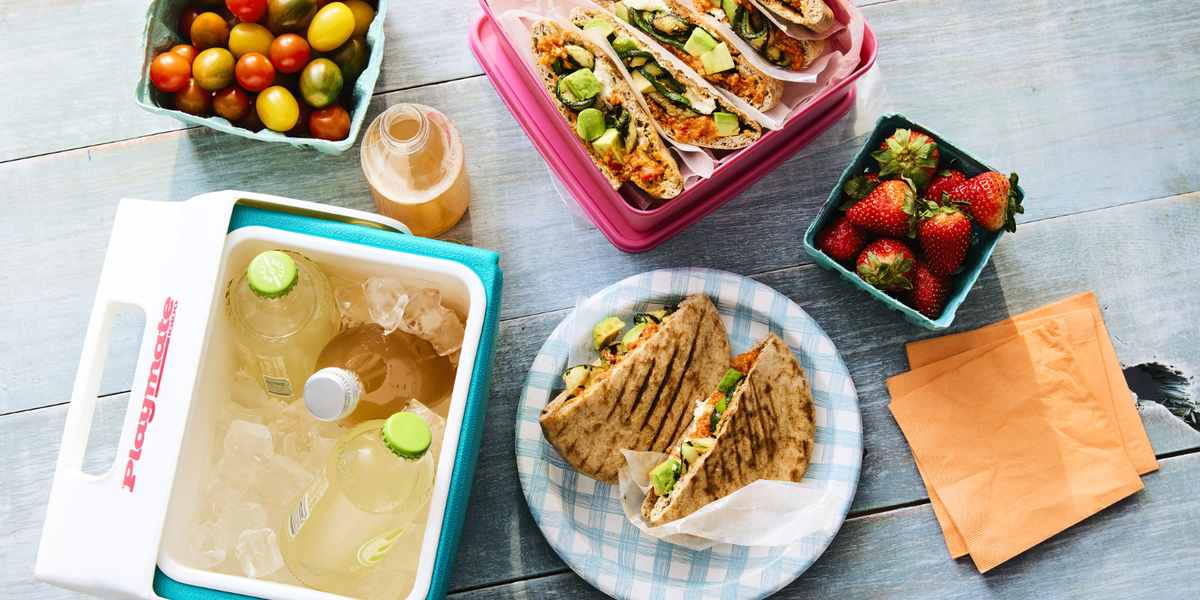 A plate and container with zucchini and romesco sandwich surrounded by fresh strawberries, tomatoes, and a cooler with refreshing beverages on a wooden grey table