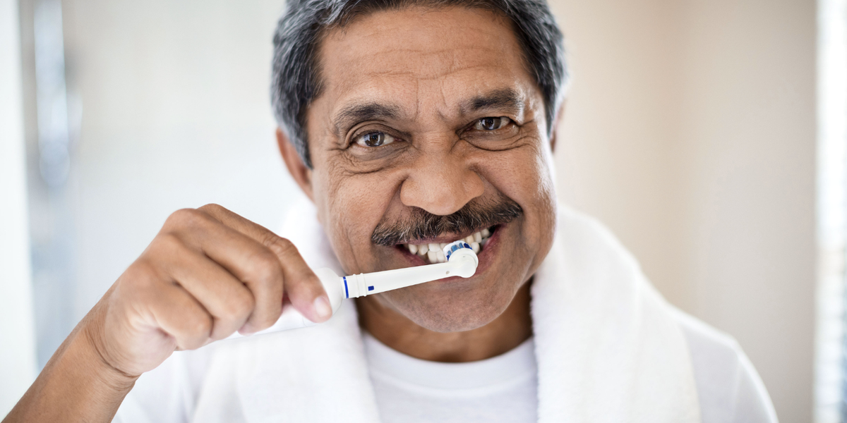 Man in a white shirt with a white towel around his neck brushing his teeth with an electric toothbrush