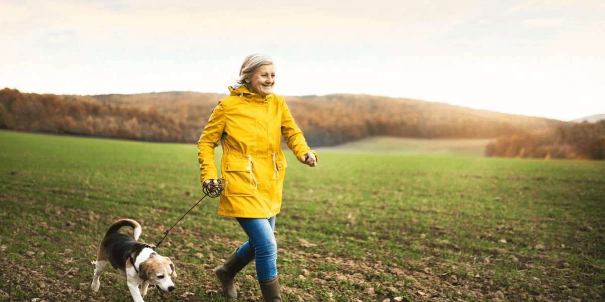Woman in yellow rain jacket walking her dog in an open field.